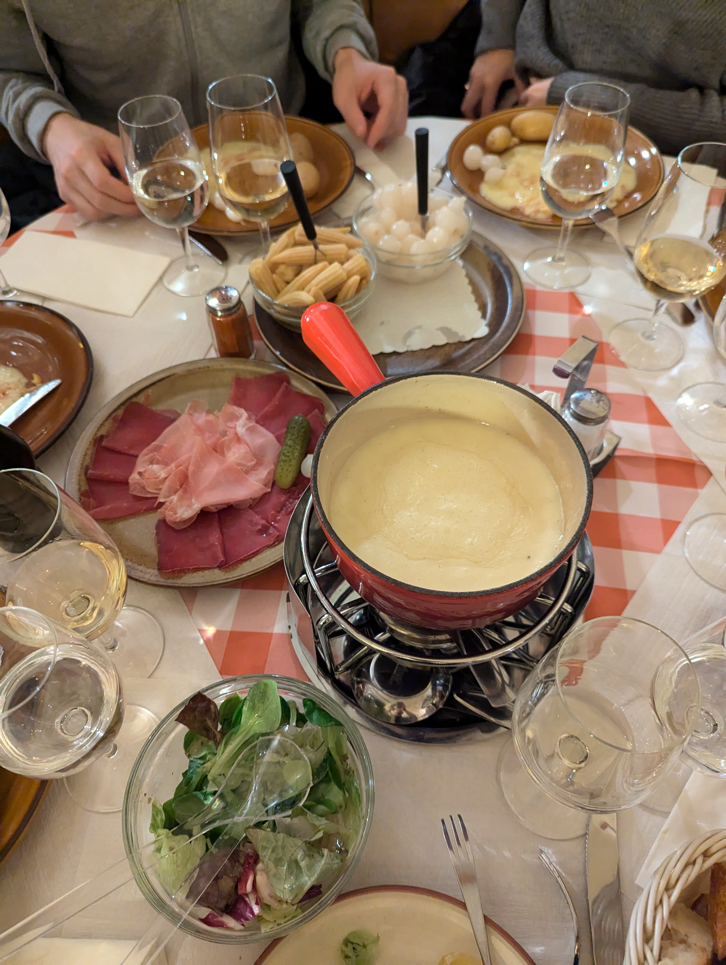 Table at Raclette Stube with wine glasses, a fondue pot, and plates of vegetables, meat, raclette, and potatoes.