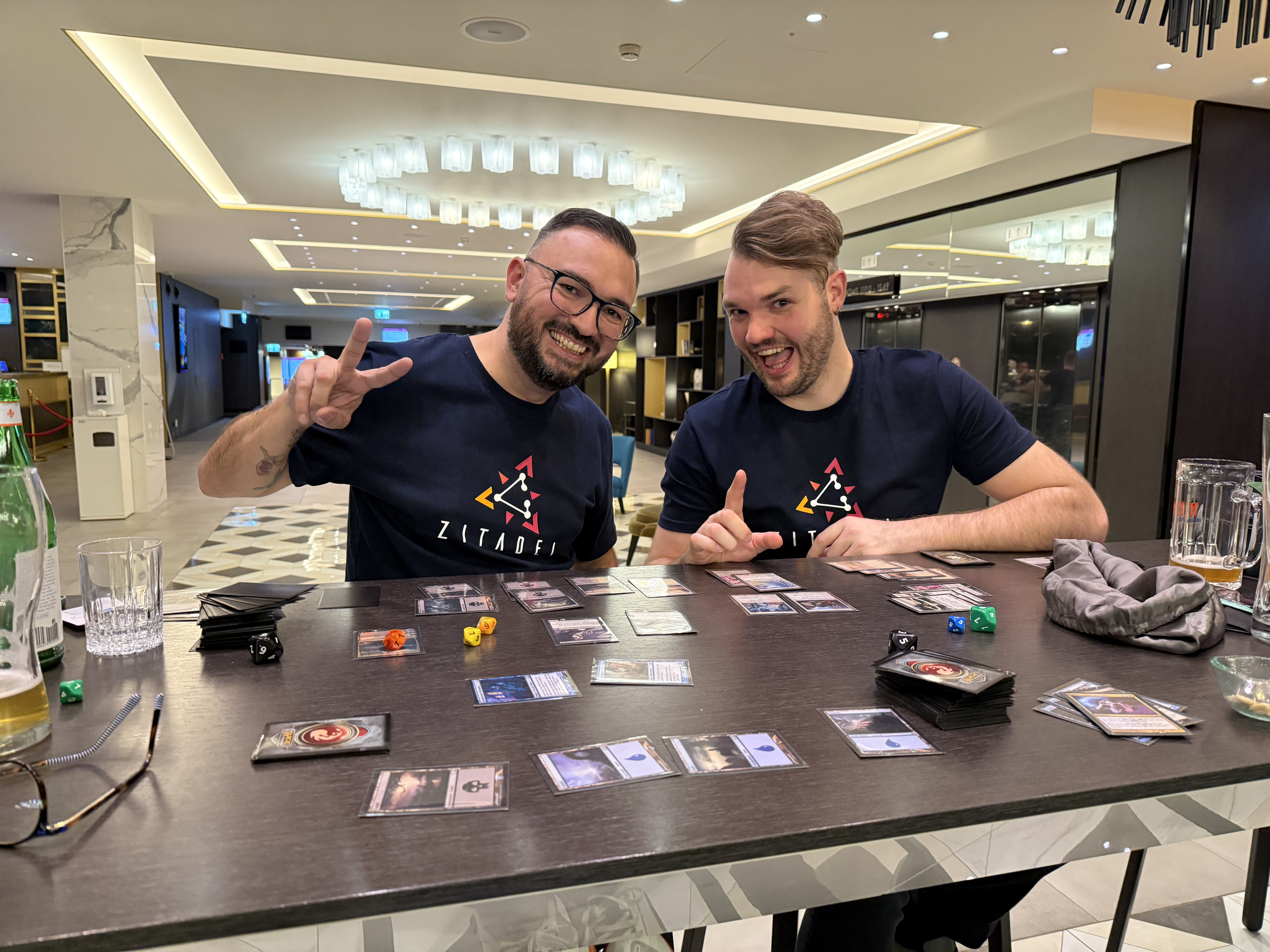 Jim and Stefan sitting in the hotel lobby playing Magic: The Gathering, with cards spread across the table and drinks nearby.